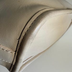 May include: Close-up of a beige leather handbag. The bag has a rounded shape with a stitched seam along the edges. The leather appears smooth and has a slight sheen. The interior of the bag is a lighter shade of beige.
