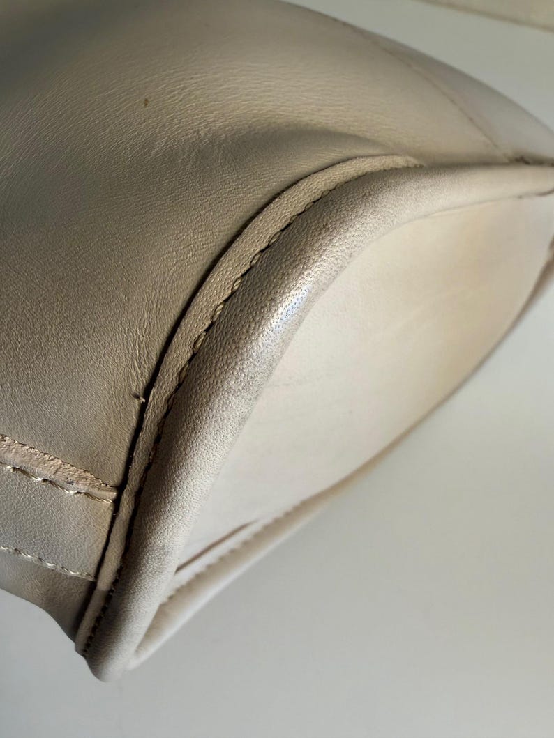 May include: Close-up of a beige leather handbag. The bag has a rounded shape with a stitched seam along the edges. The leather appears smooth and has a slight sheen. The interior of the bag is a lighter shade of beige.