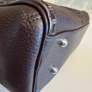 May include: Close-up of a brown leather handbag. The bag has a textured surface and silver-colored feet. The stitching and detailing are visible along the edges and corners of the bag.