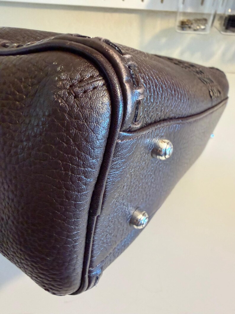 May include: Close-up of a brown leather handbag. The bag has a textured surface and silver-colored feet. The stitching and detailing are visible along the edges and corners of the bag.