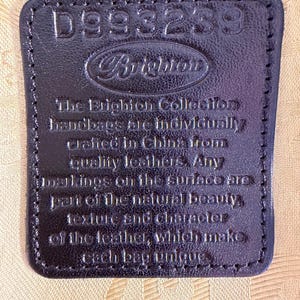 Puede incluir: Primer plano de una etiqueta de cuero negro con el texto en relieve "D993239" y "Brighton". La etiqueta tambi&eacute;n incluye texto sobre los bolsos hechos en China con cueros de calidad.