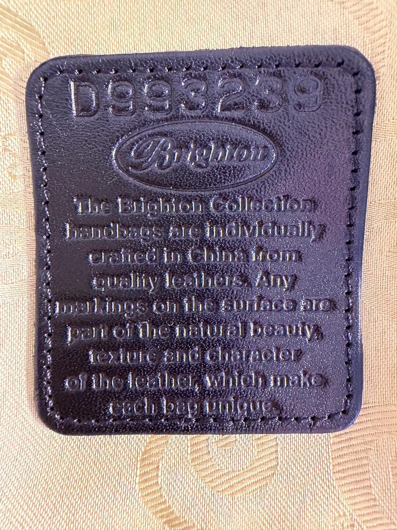 Puede incluir: Primer plano de una etiqueta de cuero negro con el texto en relieve "D993239" y "Brighton". La etiqueta tambi&eacute;n incluye texto sobre los bolsos hechos en China con cueros de calidad.
