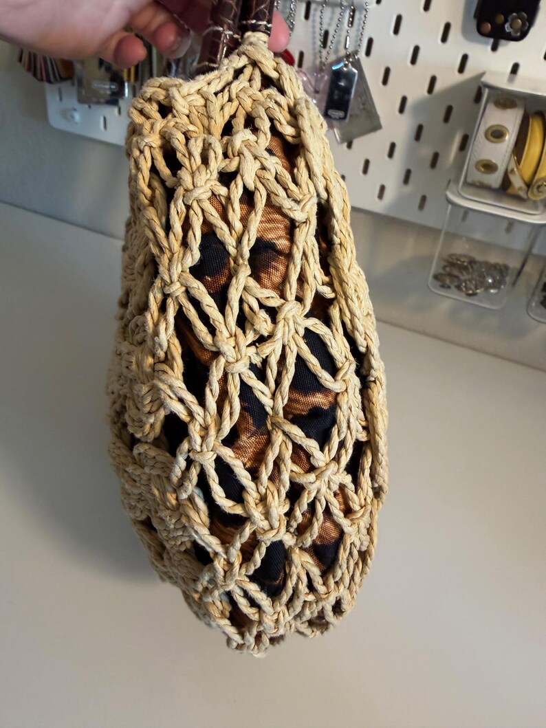 Op de afbeelding: Een beige geweven tas met een bruin lederen handvat. De tas heeft een netachtig ontwerp, dat een voering van luipaardprintstof onthult. De tas wordt door een hand vastgehouden, wat de textuur en stijl van het accessoire laat zien.