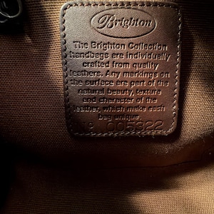 May include: Close-up of a brown leather tag inside a handbag. The tag is embossed with the Brighton logo and text that reads "The Brighton Collection handbags are individually crafted from quality leathers." The tag also has the number 005322.