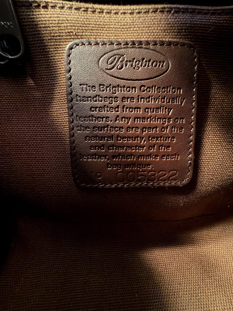 May include: Close-up of a brown leather tag inside a handbag. The tag is embossed with the Brighton logo and text that reads "The Brighton Collection handbags are individually crafted from quality leathers." The tag also has the number 005322.