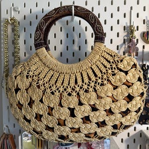 Op de afbeelding: Een beige en bruine geweven handtas met een ronde houten handgreep. De tas heeft een uniek open weefselontwerp en een patroon aan de binnenkant. De handgreep is donkerbruin met een bladvormig patroon. De tas hangt aan een wit pegboard.