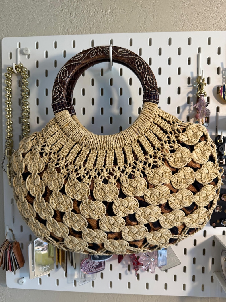 Op de afbeelding: Een beige en bruine geweven handtas met een ronde houten handgreep. De tas heeft een uniek open weefselontwerp en een patroon aan de binnenkant. De handgreep is donkerbruin met een bladvormig patroon. De tas hangt aan een wit pegboard.