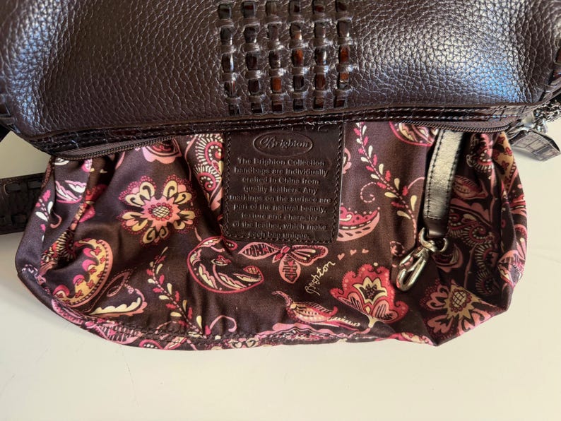 May include: A dark brown leather handbag with a woven top detail. The interior lining features a floral pattern in shades of pink, yellow, and white against a dark brown background. A leather tag is visible inside.