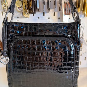 Puede incluir: Bolso negro brillante con textura de cocodrilo. El bolso tiene un bolsillo frontal con cremallera y un charm plateado en forma de coraz&oacute;n. La correa para el hombro es negra y el bolso est&aacute; colgado.
