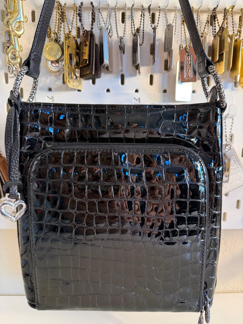 Puede incluir: Bolso negro brillante con textura de cocodrilo. El bolso tiene un bolsillo frontal con cremallera y un charm plateado en forma de coraz&oacute;n. La correa para el hombro es negra y el bolso est&aacute; colgado.