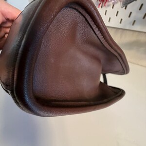 May include: A dark brown leather handbag, showcasing a curved, structured design. The bag's surface has a textured appearance, with visible stitching along the edges. The image highlights the bag's shape and material.