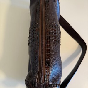 May include: A dark brown leather handbag with a woven design on the front. The bag has a zipper closure and a long, thin shoulder strap. A silver chain and decorative charm hang from the bottom.