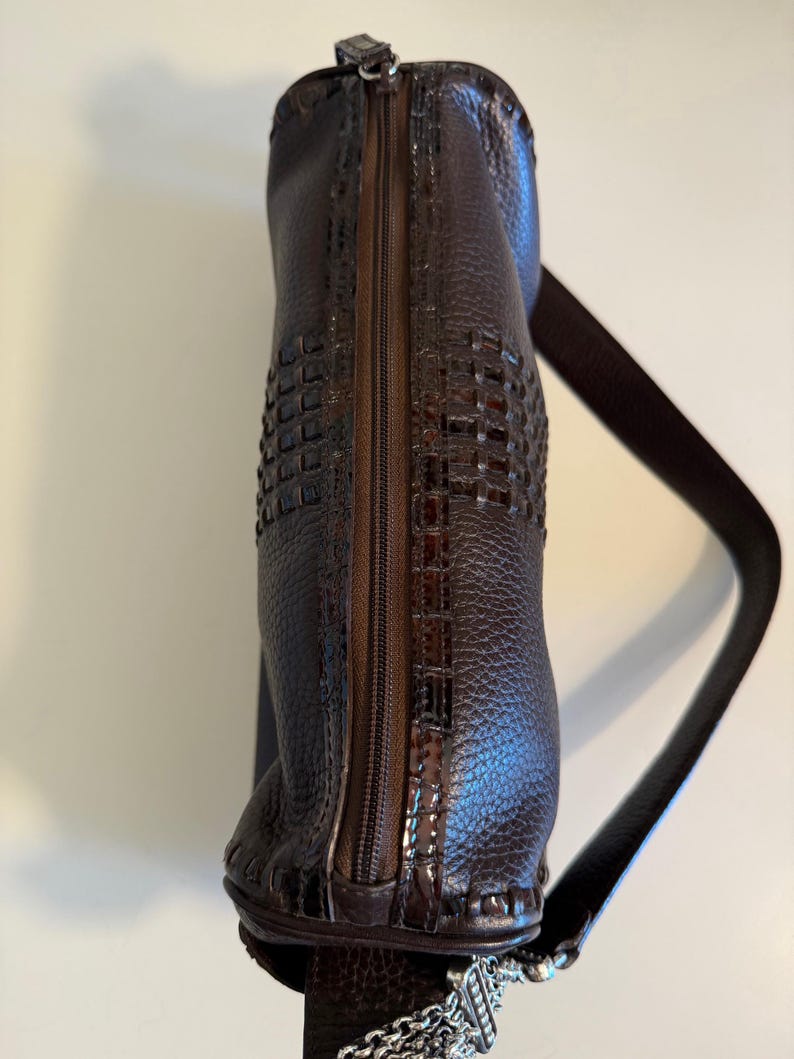 May include: A dark brown leather handbag with a woven design on the front. The bag has a zipper closure and a long, thin shoulder strap. A silver chain and decorative charm hang from the bottom.