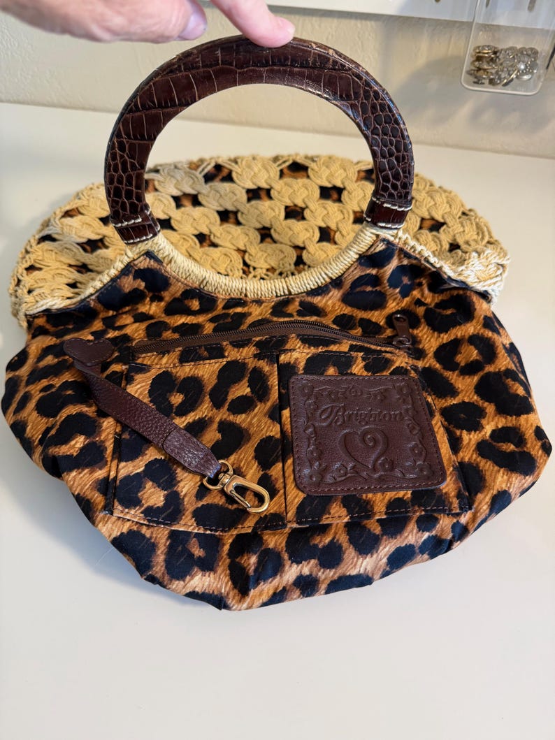 Op de afbeelding: Een handtas met luipaardprint met een bruin handvat en een geweven beige bovenkant. De tas heeft een bruin lederen accent met het woord "Brighton" in reli&euml;f. Een kleine bruine riem met een gouden sluiting is aan de tas bevestigd.