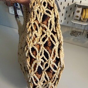 Op de afbeelding: Een beige geweven handtas met een netachtig ontwerp. De binnenstof heeft een bruine en zwarte dierenprint. De tas heeft een handvat met een donkerbruin en beige patroon.