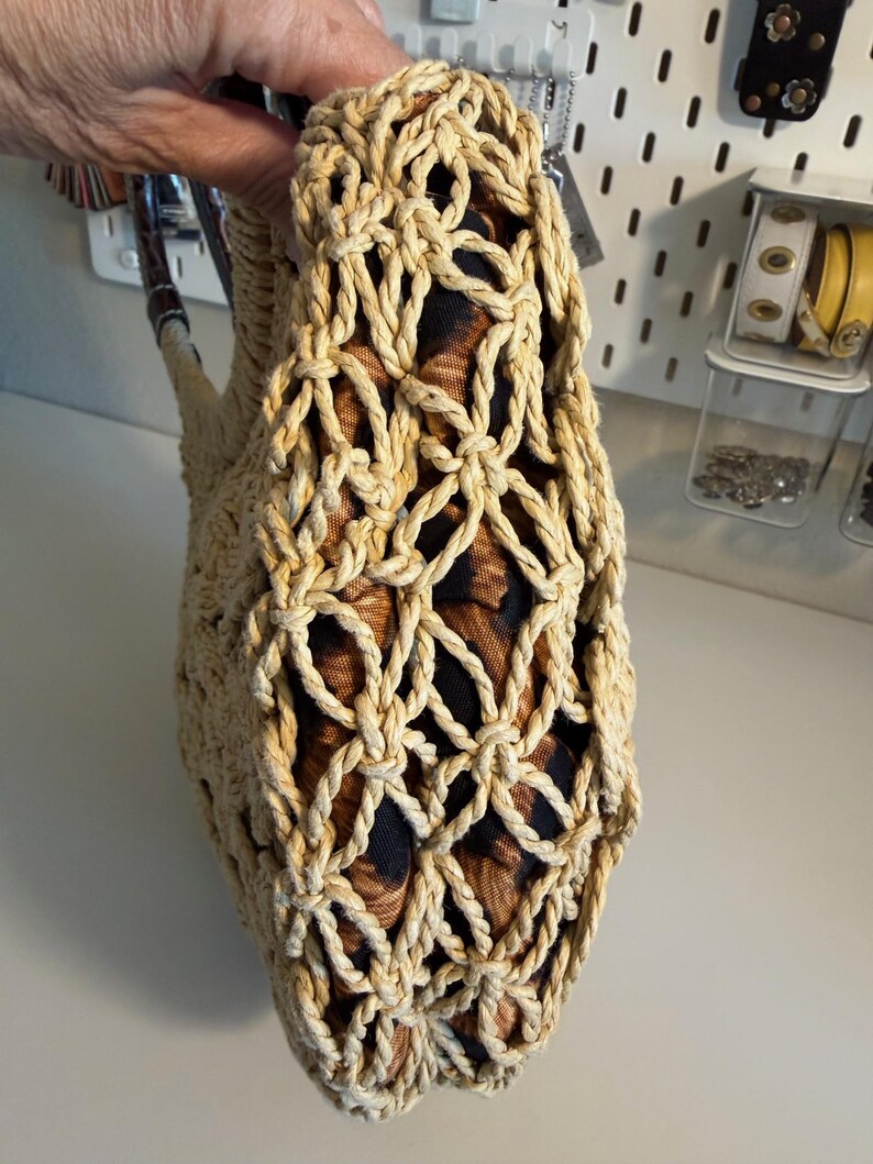 Op de afbeelding: Een beige geweven handtas met een netachtig ontwerp. De binnenstof heeft een bruine en zwarte dierenprint. De tas heeft een handvat met een donkerbruin en beige patroon.