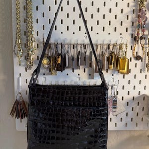 Puede incluir: Un bolso negro rectangular con un acabado brillante y texturizado, colgado de una pared perforada blanca. El bolso tiene una correa larga y delgada. Varias cadenas y etiquetas tambi&eacute;n se exhiben en la pared perforada, incluyendo cadenas doradas y plateadas.