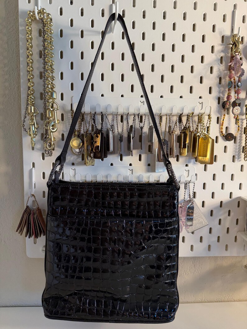 Puede incluir: Un bolso negro rectangular con un acabado brillante y texturizado, colgado de una pared perforada blanca. El bolso tiene una correa larga y delgada. Varias cadenas y etiquetas tambi&eacute;n se exhiben en la pared perforada, incluyendo cadenas doradas y plateadas.