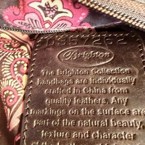 May include: Close-up of a brown leather Brighton handbag tag. The tag has the Brighton logo and text that reads, "The Brighton Collection handbags are individually crafted in China from quality leathers."
