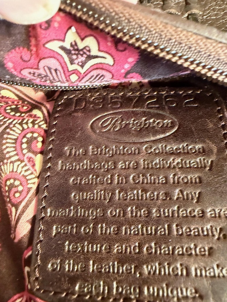 May include: Close-up of a brown leather Brighton handbag tag. The tag has the Brighton logo and text that reads, "The Brighton Collection handbags are individually crafted in China from quality leathers."