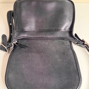 May include: Black leather shoulder bag with a flap closure. The bag has a rounded bottom and a long adjustable strap. The interior is visible, showing a suede-like texture. Silver-tone hardware accents the bag.