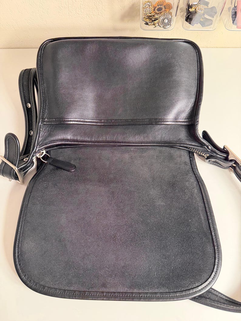 May include: Black leather shoulder bag with a flap closure. The bag has a rounded bottom and a long adjustable strap. The interior is visible, showing a suede-like texture. Silver-tone hardware accents the bag.