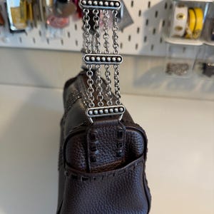 May include: A dark brown leather handbag with a textured finish. The bag features a decorative silver chain strap and stitching details. The handle is also dark brown leather. The bag is a medium size.