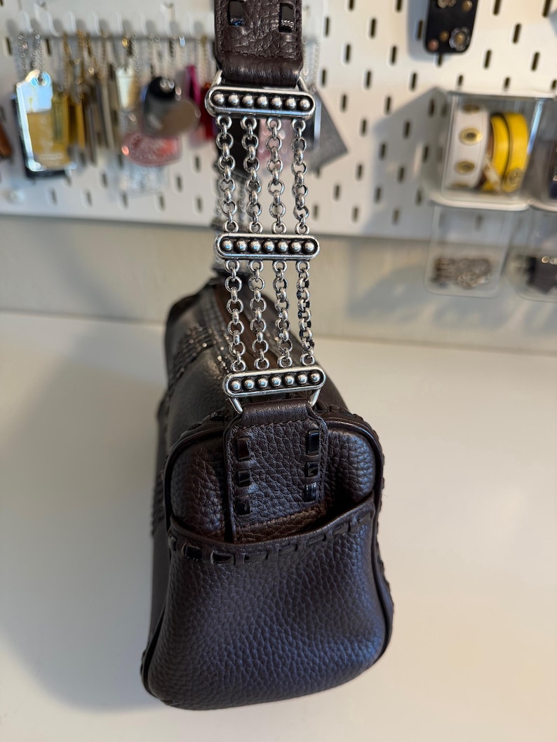 May include: A dark brown leather handbag with a textured finish. The bag features a decorative silver chain strap and stitching details. The handle is also dark brown leather. The bag is a medium size.