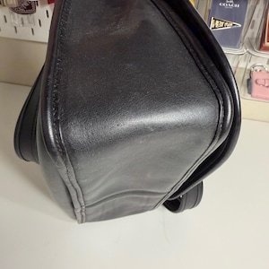 May include: A black leather shoulder bag with a flap closure. The bag has a rectangular shape with rounded corners and visible stitching along the edges. The leather appears worn.