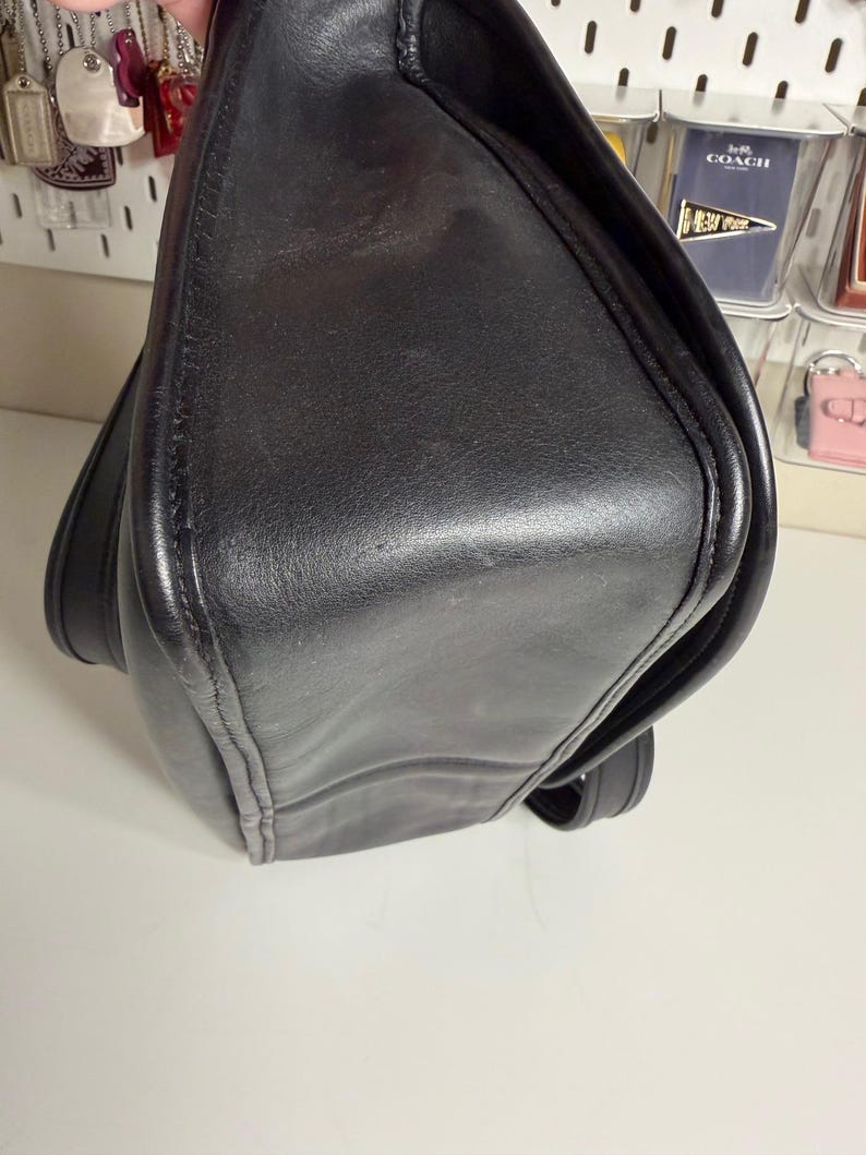 May include: A black leather shoulder bag with a flap closure. The bag has a rectangular shape with rounded corners and visible stitching along the edges. The leather appears worn.
