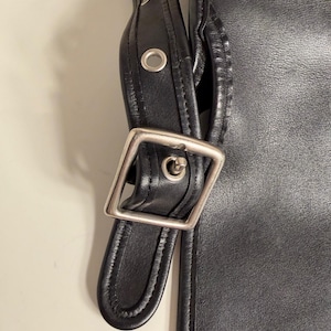 May include: Close-up of a black leather bag with a silver-colored buckle and strap. The bag has a simple, classic design with visible stitching and metal rivets. The strap is adjustable and appears durable.