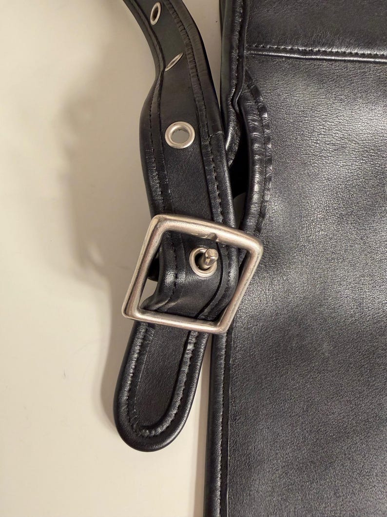 May include: Close-up of a black leather bag with a silver-colored buckle and strap. The bag has a simple, classic design with visible stitching and metal rivets. The strap is adjustable and appears durable.
