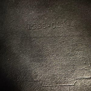 May include: Close-up of a black leather surface with the embossed text "1254-001-S". The leather has a textured appearance with subtle variations in tone. The image highlights the details of the material and the embossed markings.