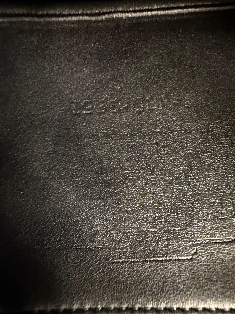 May include: Close-up of a black leather surface with the embossed text "1254-001-S". The leather has a textured appearance with subtle variations in tone. The image highlights the details of the material and the embossed markings.