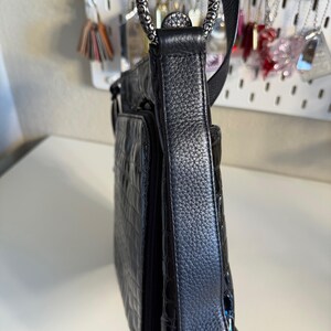 Puede incluir: Bolso bandolera de cuero negro con un patr&oacute;n texturizado similar al de un cocodrilo en el bolsillo delantero. El bolso presenta un anillo decorativo plateado y una correa negra ajustable. El bolso tiene un acabado brillante en la parte inferior.