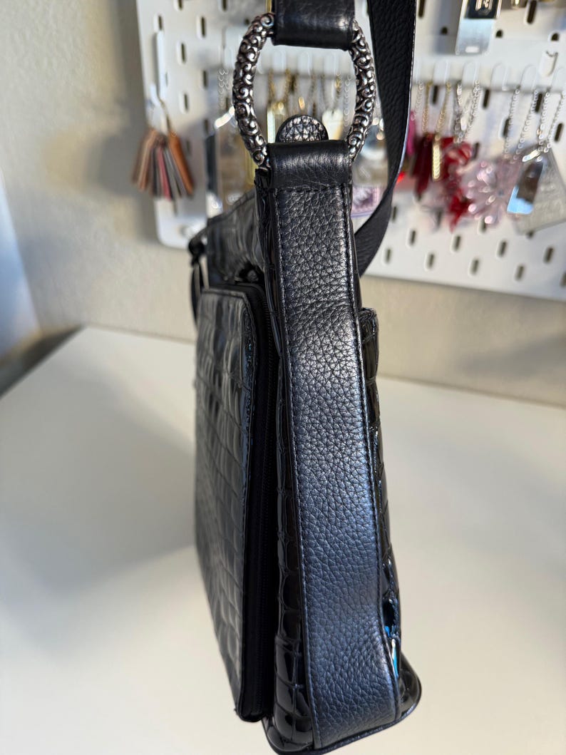 Puede incluir: Bolso bandolera de cuero negro con un patr&oacute;n texturizado similar al de un cocodrilo en el bolsillo delantero. El bolso presenta un anillo decorativo plateado y una correa negra ajustable. El bolso tiene un acabado brillante en la parte inferior.