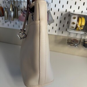 May include: Cream-colored leather tote bag with a braided brown handle. The bag features a silver heart-shaped charm and a stitched design along the side. The bag is a neutral color and appears to be made of high-quality leather.