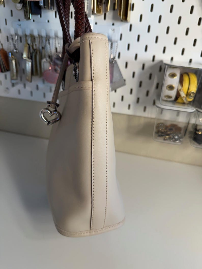 May include: Cream-colored leather tote bag with a braided brown handle. The bag features a silver heart-shaped charm and a stitched design along the side. The bag is a neutral color and appears to be made of high-quality leather.