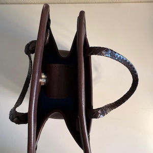 May include: A brown leather handbag with a structured design. The bag features braided handles and a silver-tone clasp. The interior is a contrasting dark blue. The bag is empty and open, showcasing its interior.