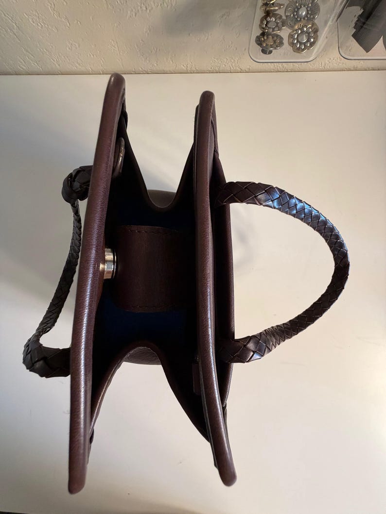May include: A brown leather handbag with a structured design. The bag features braided handles and a silver-tone clasp. The interior is a contrasting dark blue. The bag is empty and open, showcasing its interior.
