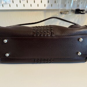 May include: A dark brown leather handbag with a woven center panel and a braided handle. The bag has silver-toned feet and stitching details. The leather has a textured appearance.