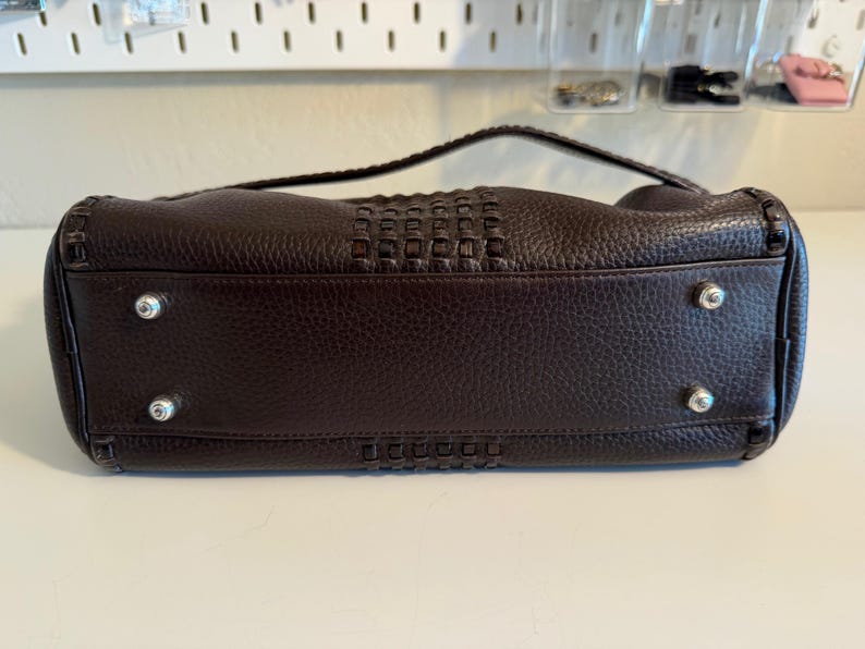 May include: A dark brown leather handbag with a woven center panel and a braided handle. The bag has silver-toned feet and stitching details. The leather has a textured appearance.