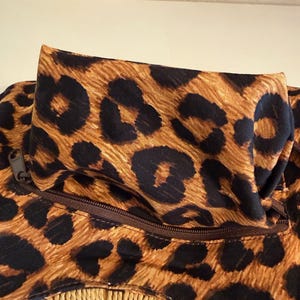 Op de afbeelding: Close-up van een tas met een luipaardprint in bruin en zwart. De tas heeft een rits en een geweven, beige franje aan de onderkant. De tas is gemaakt van stof.