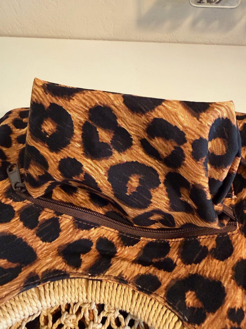 Op de afbeelding: Close-up van een tas met een luipaardprint in bruin en zwart. De tas heeft een rits en een geweven, beige franje aan de onderkant. De tas is gemaakt van stof.
