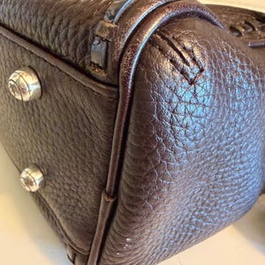May include: Close-up of a dark brown leather handbag. The bag has a textured surface and silver-colored metal studs on the bottom. The stitching and trim are visible, highlighting the craftsmanship of the bag.