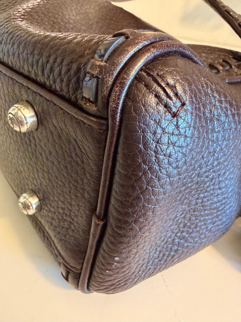 May include: Close-up of a dark brown leather handbag. The bag has a textured surface and silver-colored metal studs on the bottom. The stitching and trim are visible, highlighting the craftsmanship of the bag.