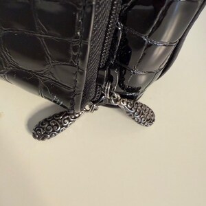 Puede incluir: Primer plano de un bolso negro brillante y texturizado con cremallera. El tirador de la cremallera tiene un dise&ntilde;o ornamentado de color plateado. La superficie del bolso tiene un patr&oacute;n similar al de un cocodrilo.