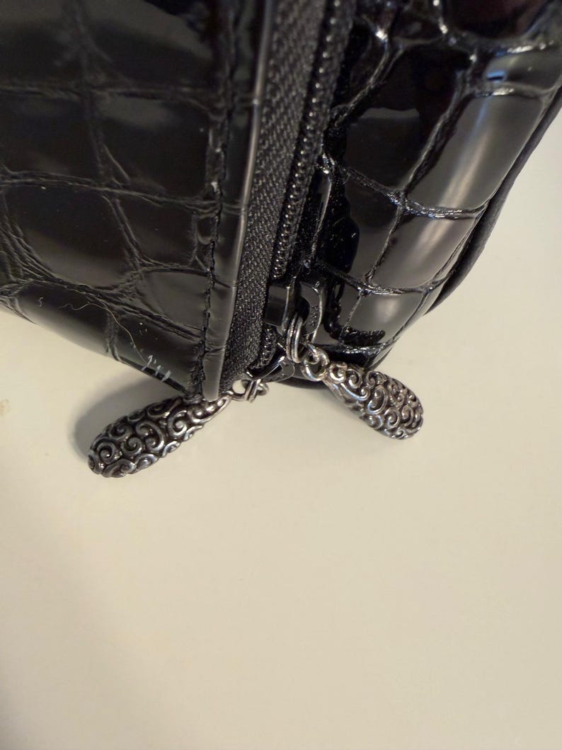 Puede incluir: Primer plano de un bolso negro brillante y texturizado con cremallera. El tirador de la cremallera tiene un dise&ntilde;o ornamentado de color plateado. La superficie del bolso tiene un patr&oacute;n similar al de un cocodrilo.