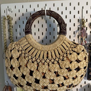 Op de afbeelding: Een beige en bruine handtas met een ronde handgreep. De tas heeft een geweven ontwerp met een luipaardprint. De handgreep is bruin met een bladerpatroon. De tas hangt aan een wit pegboard.