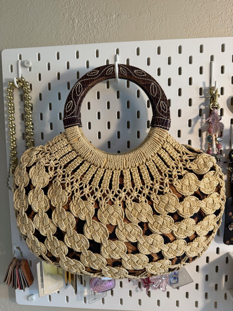 Op de afbeelding: Een beige en bruine handtas met een ronde handgreep. De tas heeft een geweven ontwerp met een luipaardprint. De handgreep is bruin met een bladerpatroon. De tas hangt aan een wit pegboard.